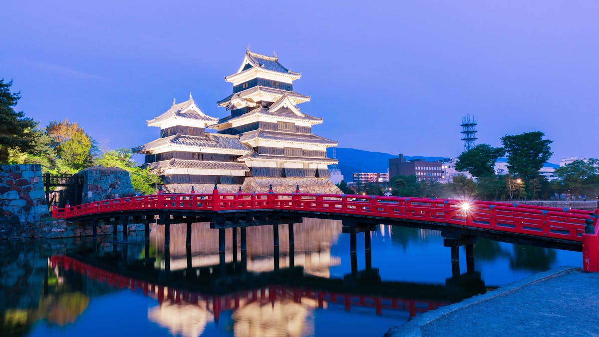 An enchanting evening shot of the historic Matsumoto Castle, with its striking black-and-white facade reflecting in the calm, surrounding water. A vivid red bridge crosses the serene moat, inviting visitors to explore the magnificent structure